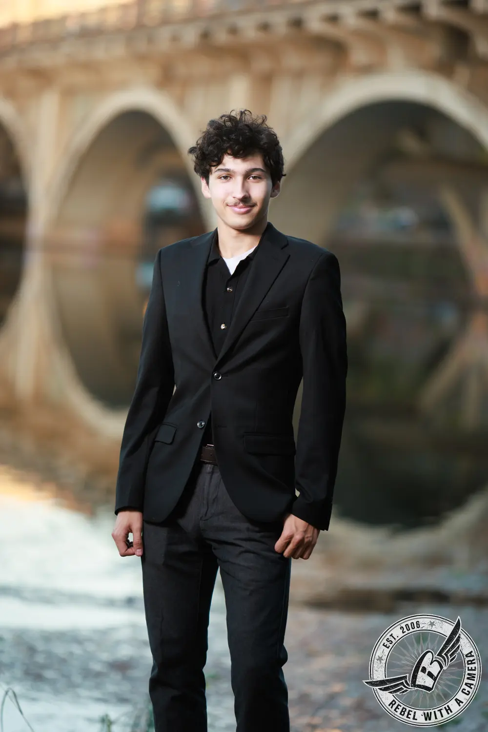 Nathan standing in a black blazer beside the water with a blurred arched bridge behind him.