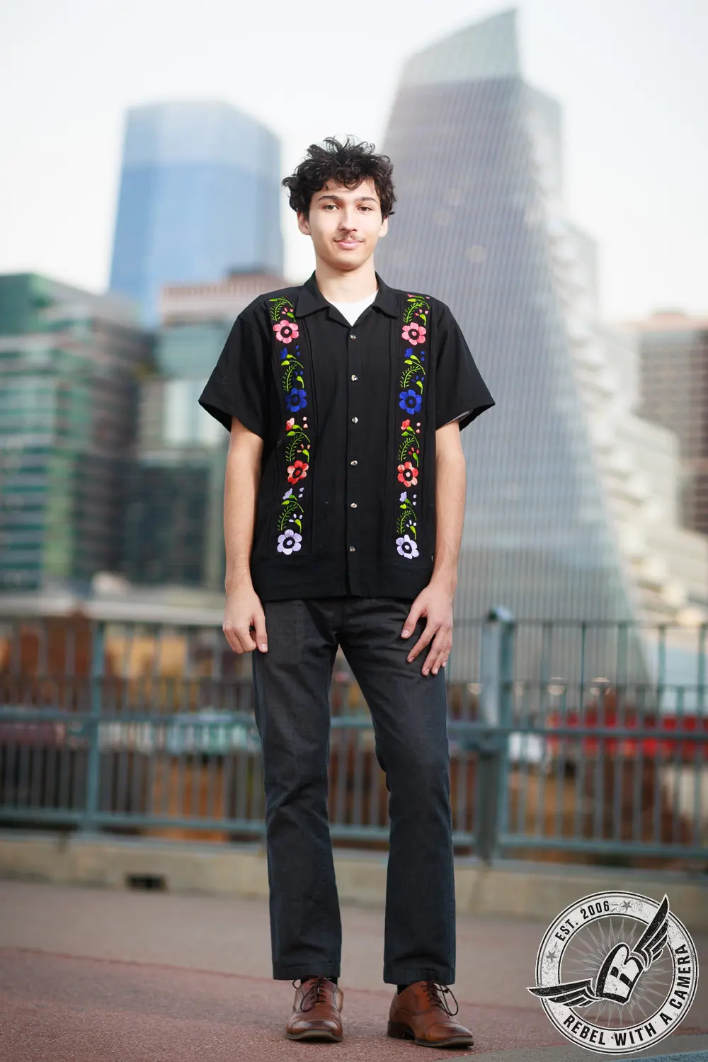 Full-length portrait of Nathan standing in front of blurred downtown buildings, wearing a black shirt with colorful floral embroidery.