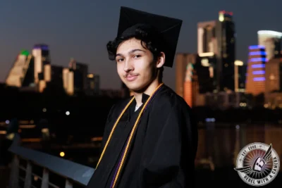 Nathan graduation portrait in a black cap and gown with the city skyline in Austin, TX.