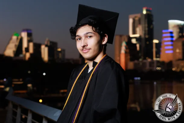 Nathan graduation portrait in a black cap and gown with the city skyline in Austin, TX.