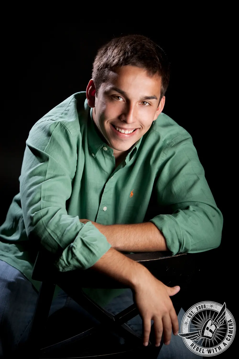 Nick wearing a green button-down shirt and leaning on a black chair against a black background.
