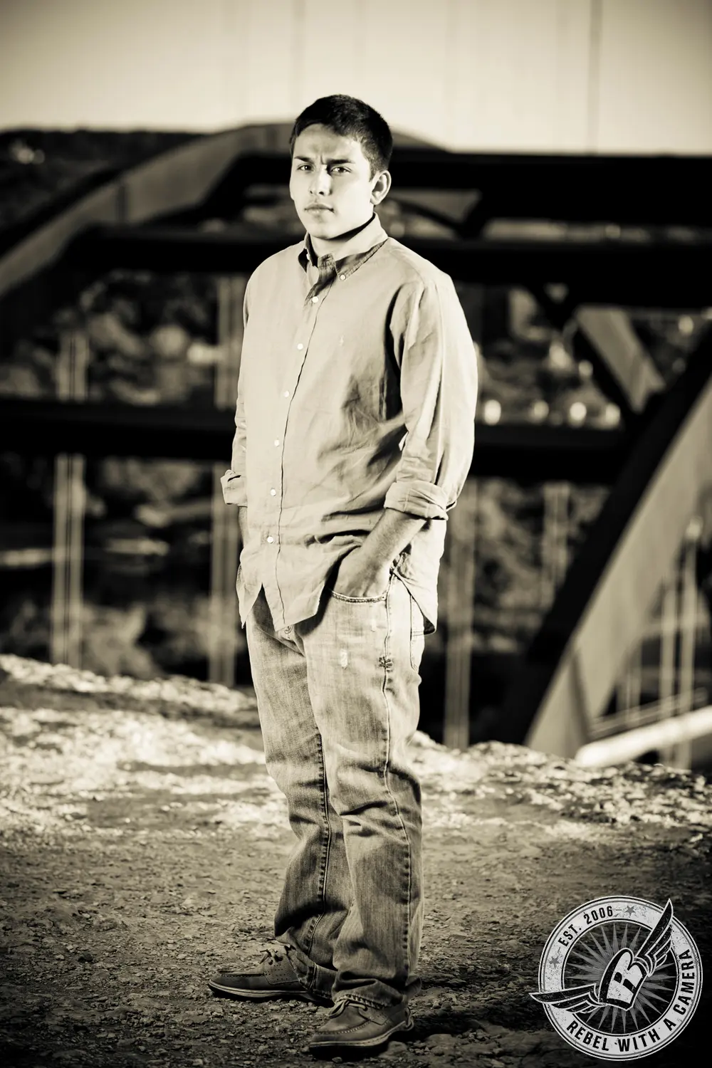 Sepia-toned full-length portrait of Nick in a button-down shirt and jeans standing near bridge supports.