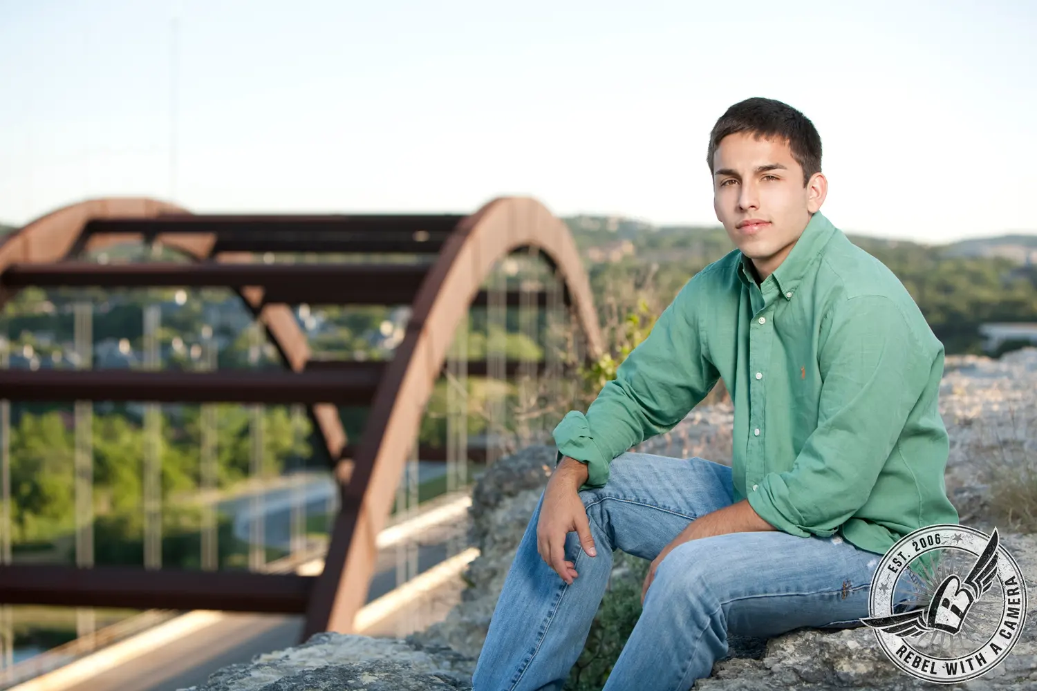 Nick seated on a rock in a green shirt and jeans with Pennybacker Bridge behind him.