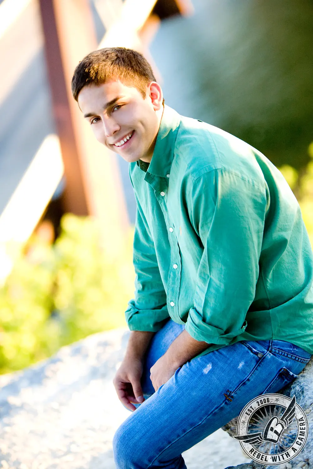 Nick, wearing a green button-down shirt and jeans, seated on a rock outdoors and smiling toward the camera.