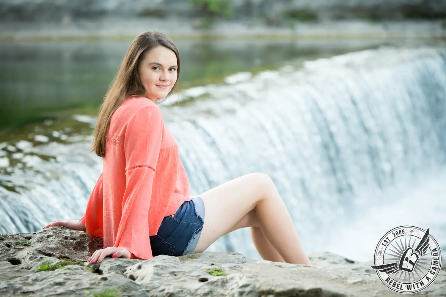 Nicole seated on a rock beside a small waterfall at Brushy Creek, wearing a coral blouse and denim shorts.