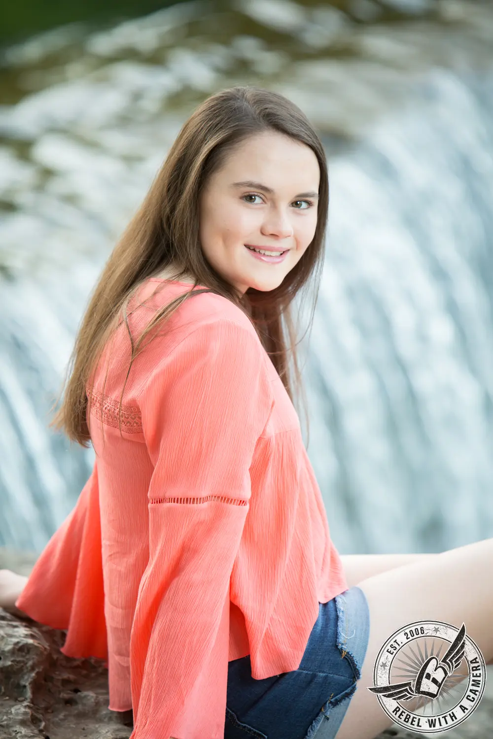 Nicole seated on a rock near flowing water, wearing a coral blouse and denim shorts.