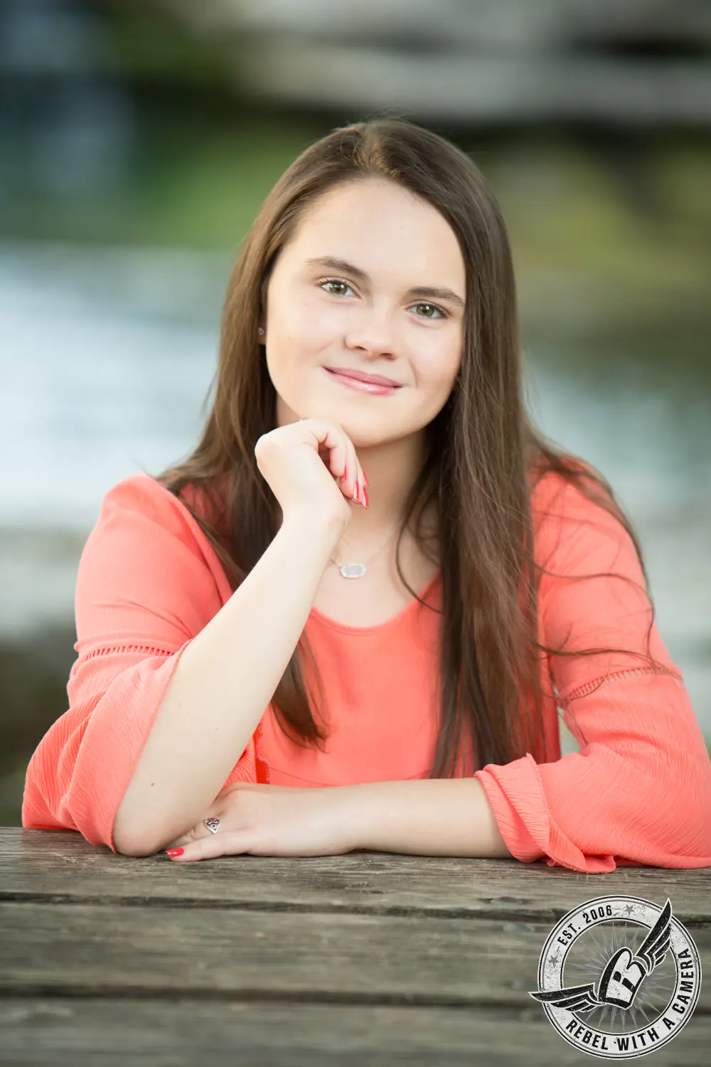 Nicole in a coral blouse resting her chin on her hand at a wooden railing, with softly blurred water and greenery behind her.