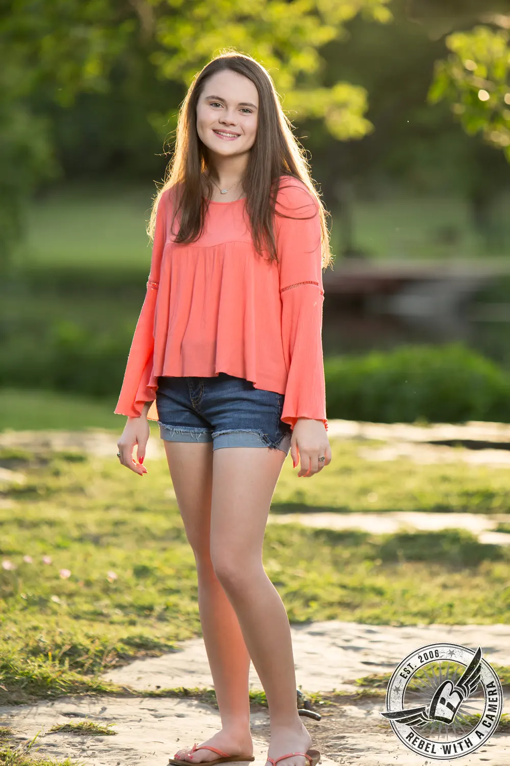 Full-length senior portrait of Nicole standing on a sunlit path in a coral blouse and denim shorts, with green trees behind her.