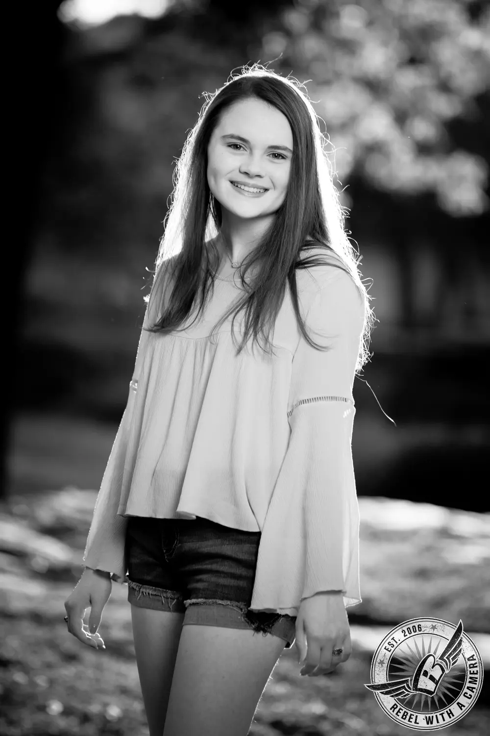 Black-and-white portrait of Nicole standing outdoors near Brushy Creek, wearing a flowing blouse and denim shorts.