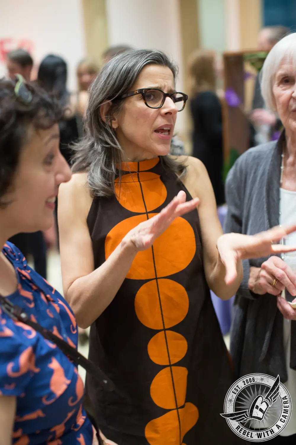 Nina Katchadourian, wearing a brown dress with large orange circles, gestures while speaking among guests indoors.