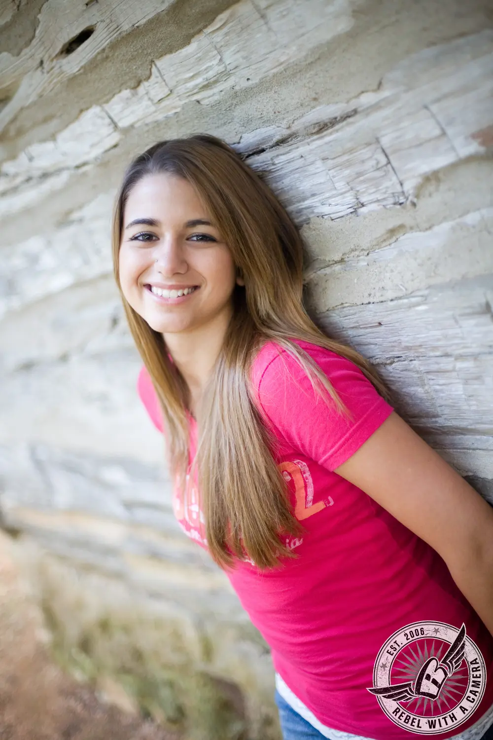 Ruby smiling while leaning against a textured pale stone wall in a bright pink shirt.