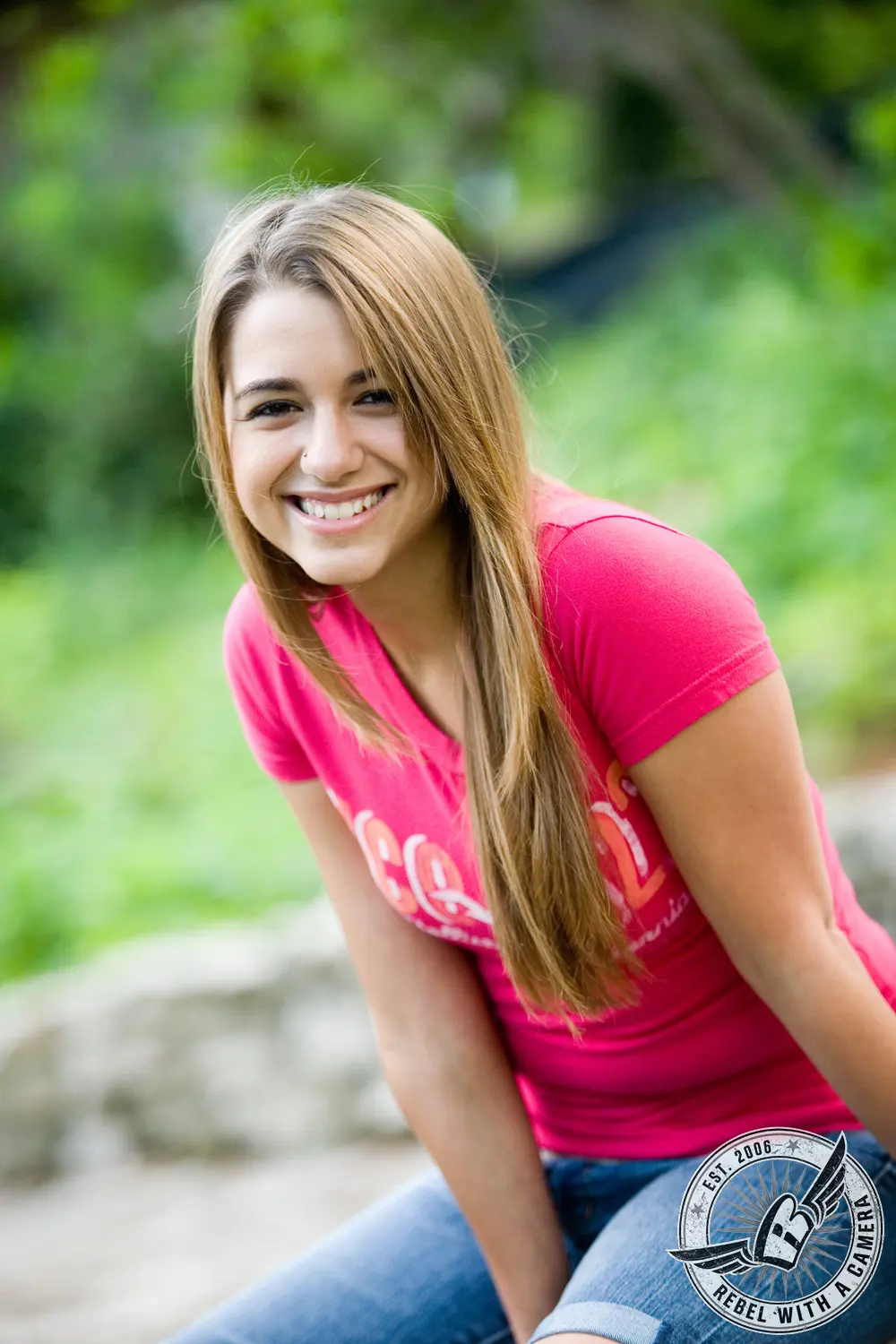 Ruby smiling in a pink T-shirt and jeans against a softly blurred green garden background.