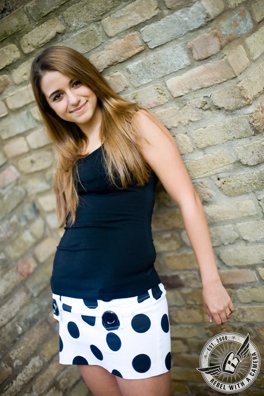 Ruby standing beside a textured stone wall in a black top and white polka-dot skirt.