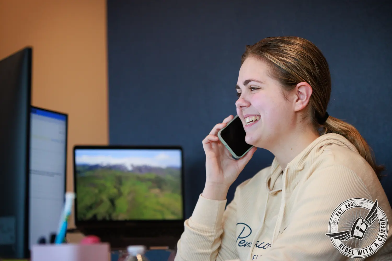 Office employee holding a smartphone beside two computer monitors.