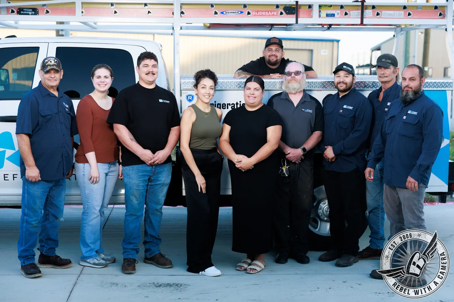 Nine Tri-Point Refrigeration employees pose in front of a branded service truck beneath mounted ladders.